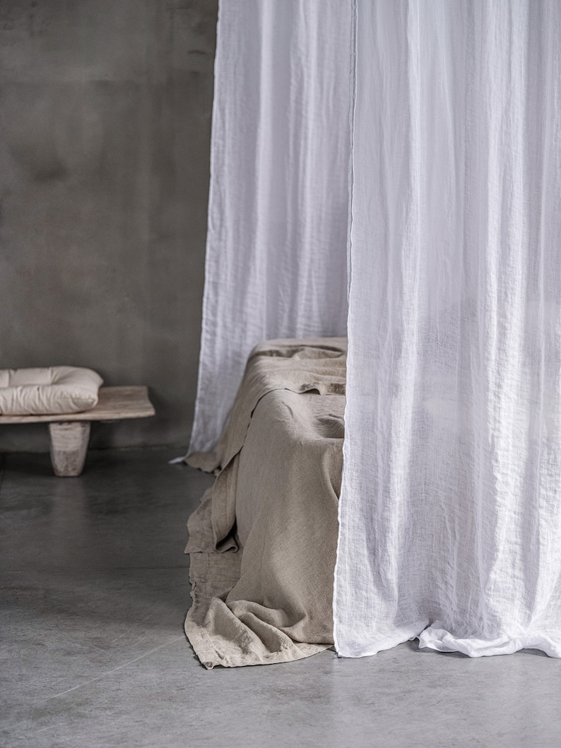 May include: A white linen canopy draped over a bed with a light brown linen duvet. The bed is in a room with a concrete floor and a grey wall.