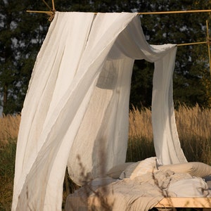 May include: A white linen canopy draped over a bed frame made of natural wood branches. The bed is covered in a beige linen blanket and pillows. The bed is set up outdoors in a field of tall grass.