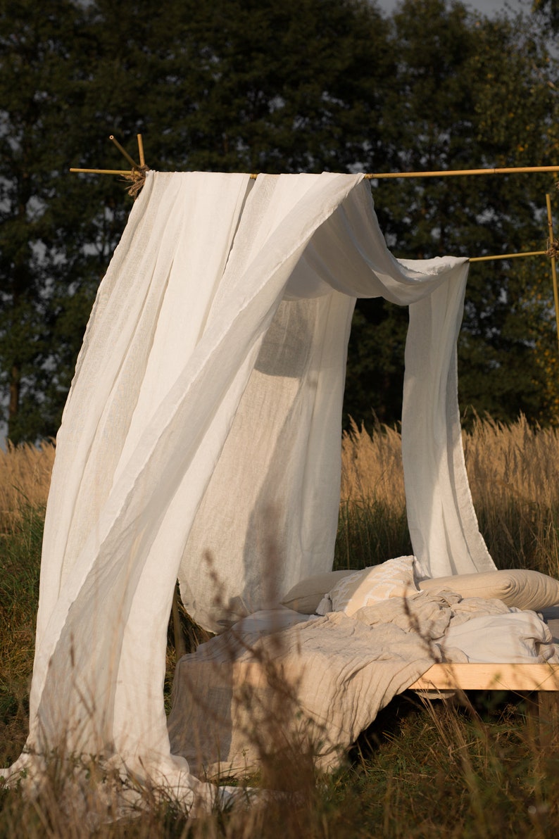 May include: A white linen canopy draped over a bed frame made of natural wood branches. The bed is covered in a beige linen blanket and pillows. The bed is set up outdoors in a field of tall grass.