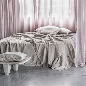 May include: A light brown linen bedspread with a white pillow on a bed with sheer pink curtains.