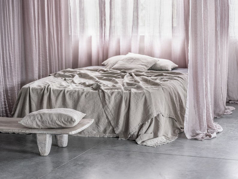 May include: A light brown linen bedspread with a white pillow on a bed with sheer pink curtains.