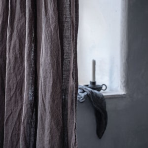 K&ouml;nnte beinhalten: Eine Nahaufnahme eines braunen Leinenvorhangs mit einer strukturierten Webart. Der Vorhang ist teilweise gezogen und gibt den Blick auf ein Fenster mit wei&szlig;em Rahmen und einen Blick auf einen Kerzenhalter frei.