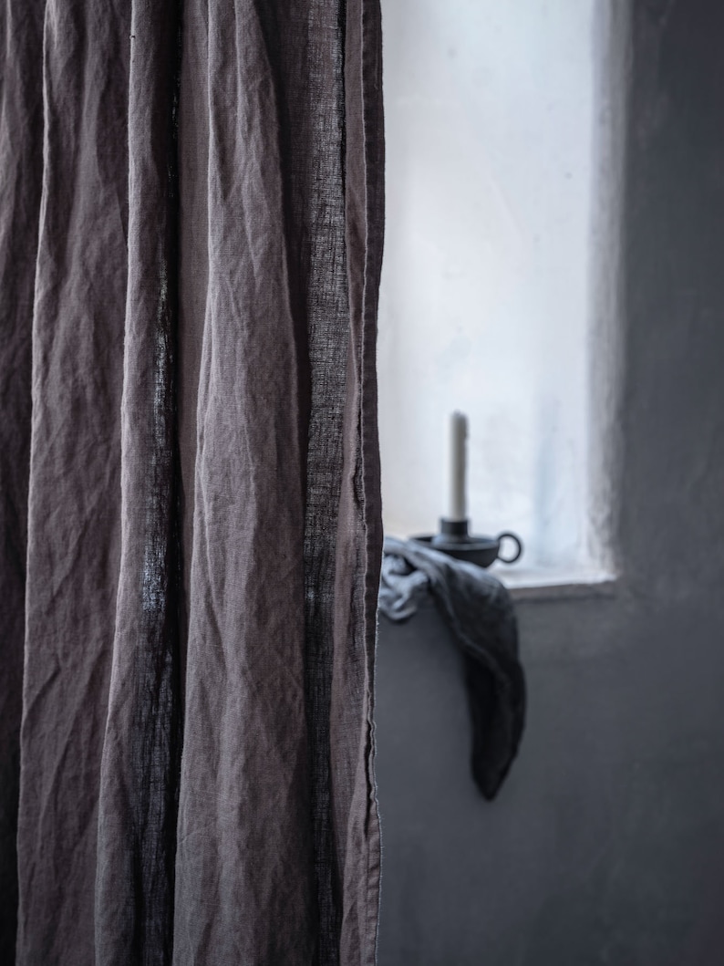 K&ouml;nnte beinhalten: Eine Nahaufnahme eines braunen Leinenvorhangs mit einer strukturierten Webart. Der Vorhang ist teilweise gezogen und gibt den Blick auf ein Fenster mit wei&szlig;em Rahmen und einen Blick auf einen Kerzenhalter frei.