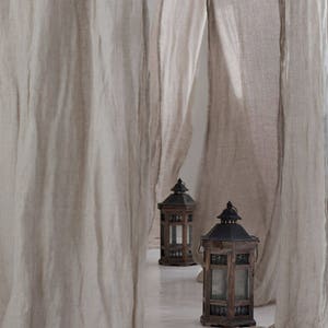 Op de afbeelding: Twee houten lantaarns met zwarte metalen accenten en glazen panelen staan op een wit oppervlak omringd door vloeiende beige linnen stof.