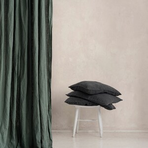 May include: A stack of three dark gray linen pillows on a white wooden stool in front of a green linen curtain.