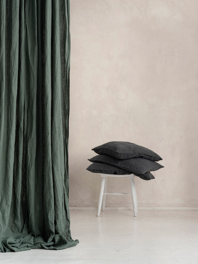 May include: A stack of three dark gray linen pillows on a white wooden stool in front of a green linen curtain.