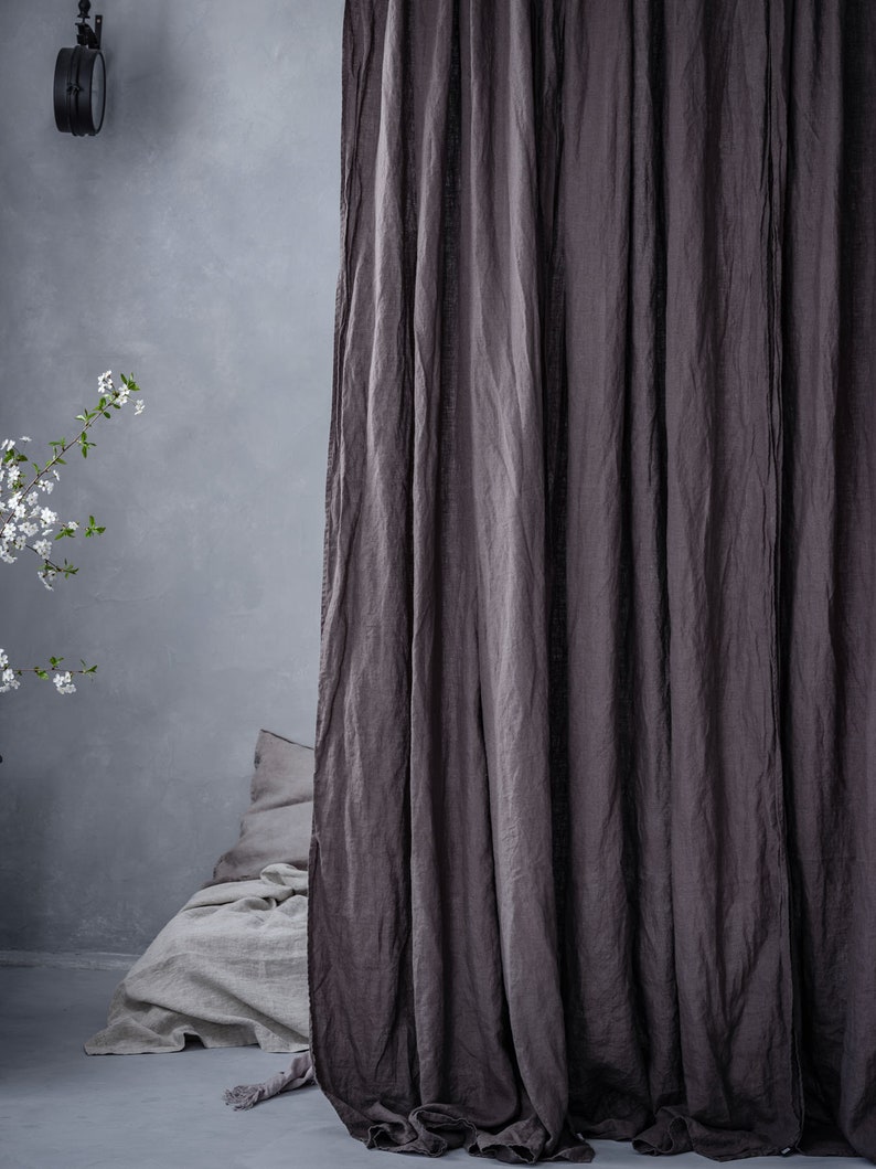 K&ouml;nnte beinhalten: Ein brauner Leinenvorhang h&auml;ngt vor einem Bett mit einer wei&szlig;en Leinenbettw&auml;sche und einem Kissen. Der Vorhang ist aus einem weichen, strukturierten Stoff und hat einen nat&uuml;rlichen, rustikalen Look.