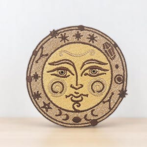 May include: Embroidered patch featuring a stylised sun face with a brown outline and gold details. The sun is surrounded by a circle of stars and crescent moons.