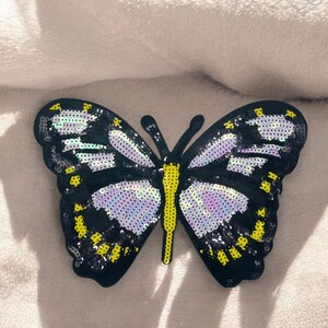 Può includere: Un cerotto di paillettes a forma di farfalla nero e giallo con ali iridescenti. Il cerotto è su uno sfondo di tessuto bianco.