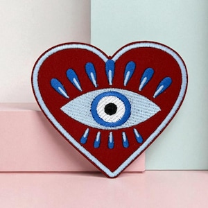 Puede incluir: Un parche bordado en forma de corazón de color rojo intenso, con un diseño de ojo azul claro con una pupila negra. El ojo está rodeado de formas de lágrima azules y detalles blancos. El parche tiene un borde blanco.