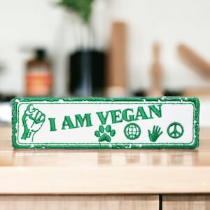 Könnte beinhalten: Weißer rechteckiger Patch mit grünem gesticktem Text "I AM VEGAN" und grünen gestickten Symbolen, darunter eine Faust, ein Pfotenabdruck, ein Globus, eine Hand und ein Friedenszeichen.