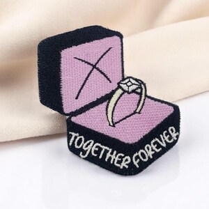Puede incluir: Parche bordado con un anillo en una caja. El anillo tiene una piedra en forma de diamante. La caja es negra con el interior rosa y las palabras "TOGETHER FOREVER" bordadas en blanco.