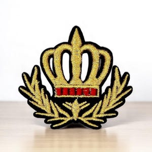 Peut inclure: Écusson brodé représentant une couronne dorée avec des détails rouges et des feuilles de laurier. Le patch a une bordure noire. Le design est détaillé et destiné à être appliqué sur des vêtements ou des accessoires. Le patch est de haute qualité.