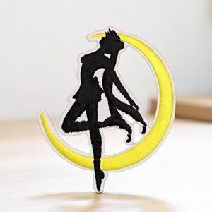 May include: Black and white embroidered patch of a silhouette of a woman standing on a yellow crescent moon.