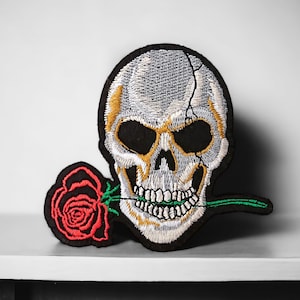 Peut inclure: Patch brodé représentant un crâne blanc avec des orbites noires et un contour doré, tenant une rose rouge dans ses dents. La rose a des feuilles vertes et une tige.