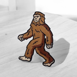 Peut inclure: Écusson brodé représentant un Bigfoot brun en marche. Le patch présente des coutures détaillées, mettant en valeur la fourrure et les traits du visage de la créature. Le design est posé sur un fond neutre, soulignant les couleurs et la forme du patch.