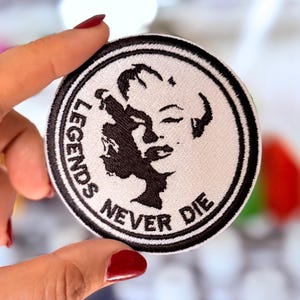 Può includere: Una toppa ricamata bianca e nera con una silhouette di un viso femminile e il testo "Legends Never Die".