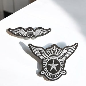 Peut inclure: Deux écussons brodés sur le thème militaire. L'un présente un bouclier ailé, l'autre une étoile, une couronne et des ailes. Les deux écussons ont un fond noir avec des coutures blanches.