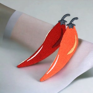 Könnte beinhalten: Zwei rote Chili-Pfeffer-Stickereien mit schwarzen Nähten. Die Paprika sind kreuzförmig angeordnet.