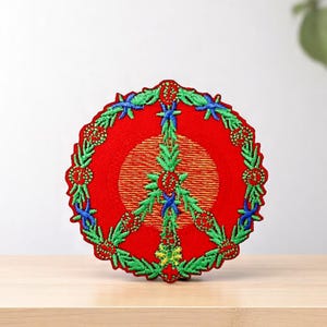May include: Embroidered peace sign patch with a red background and a circular design. The peace symbol is green with blue accents, surrounded by a green wreath with red berries. The patch is on a wooden surface.