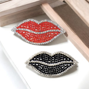 Può includere: Due spille a forma di labbra in strass, una rossa e una nera. Le spille sono fatte di strass e hanno un retro in metallo argentato.