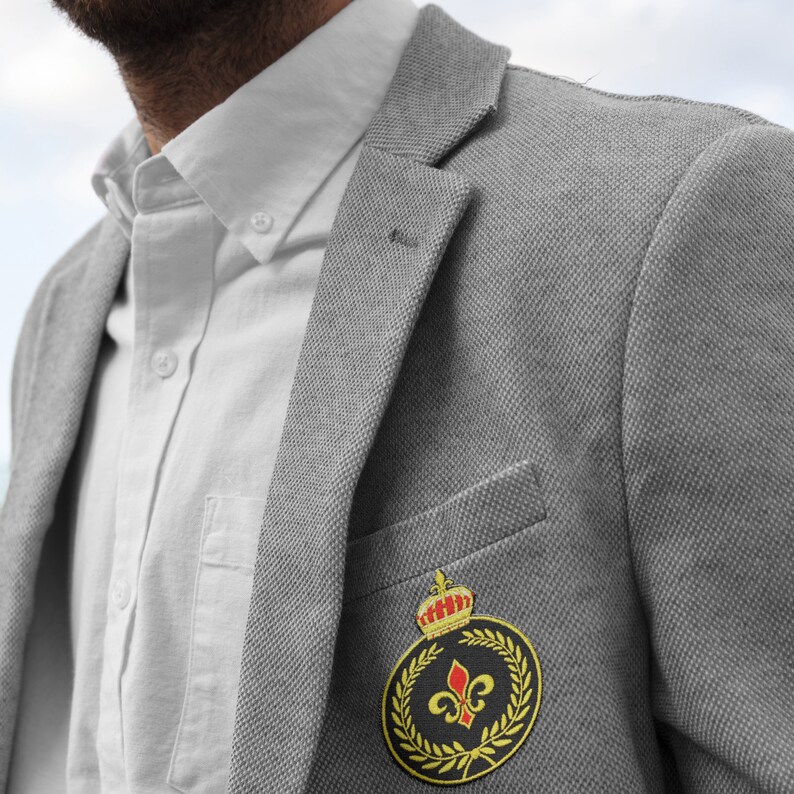 May include: A gray blazer with a gold and black embroidered patch featuring a crown, a fleur-de-lis, and a wreath of leaves.