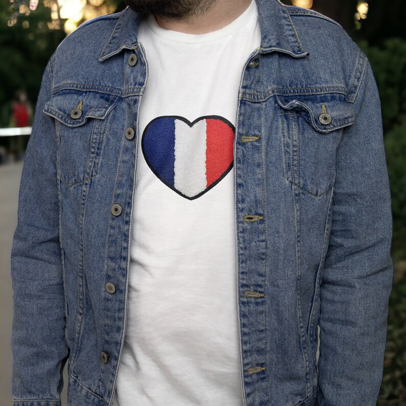 Puede incluir: Una camiseta blanca con un dise&ntilde;o en forma de coraz&oacute;n azul, blanco y rojo. La camiseta se lleva debajo de una chaqueta vaquera azul.