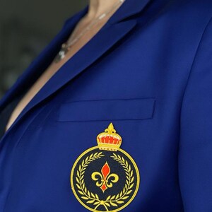 May include: A royal blue blazer with a gold and black embroidered patch featuring a crown, a fleur-de-lis, and a laurel wreath.