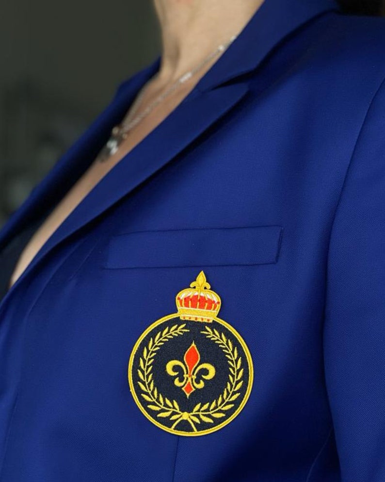 May include: A royal blue blazer with a gold and black embroidered patch featuring a crown, a fleur-de-lis, and a laurel wreath.