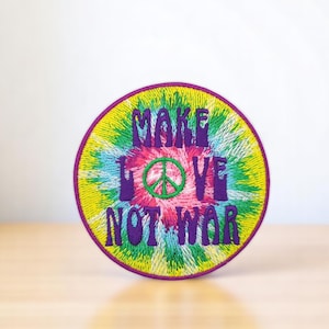 Puede incluir: Un parche redondo de tie-dye con el texto "Make Love Not War" en letras moradas. Un símbolo de paz verde está centrado en el texto.