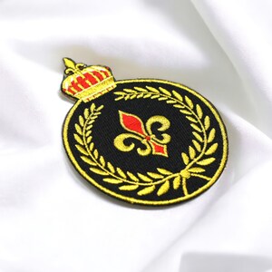May include: A gold embroidered patch with a black background featuring a red and gold fleur-de-lis surrounded by a gold laurel wreath and a gold crown at the top.