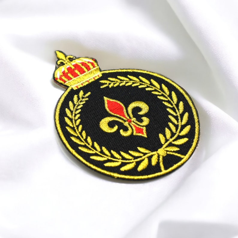 May include: A gold embroidered patch with a black background featuring a red and gold fleur-de-lis surrounded by a gold laurel wreath and a gold crown at the top.