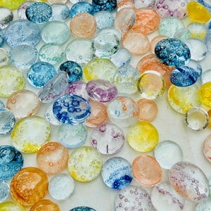 May include: A collection of numerous circular glass cabochons in various translucent colors including blue, yellow, orange, and clear. Each piece features a smooth, rounded top surface and contains small internal air bubbles throughout the glass.