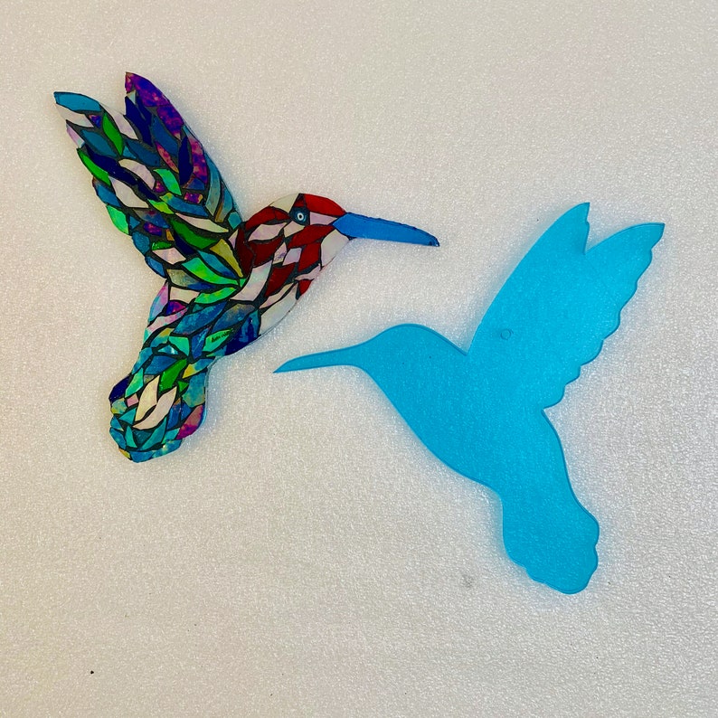 May include: Two hummingbird silhouettes, one made of colourful mosaic glass and the other a solid blue, are displayed against a white background.