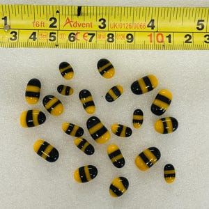 May include: A selection of small, oval glass beads featuring a yellow and black striped pattern. The beads are arranged on a white surface. A yellow measuring tape is in the background.