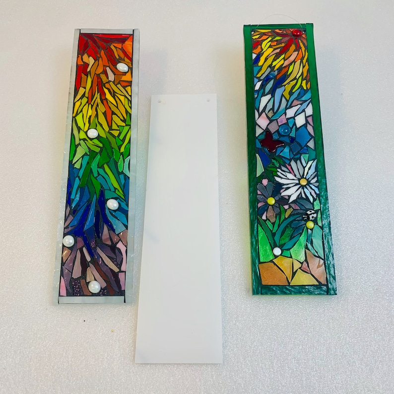May include: Two colourful mosaic glass suncatchers, one with a rainbow design and the other with a floral design. Both suncatchers are rectangular and have a green border.