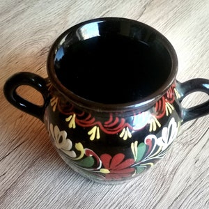 May include: A black ceramic pitcher with two handles and a floral design in red, yellow, white, and green.