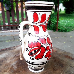 May include: A white ceramic pitcher with a red and black floral design. The pitcher has a handle and a spout.