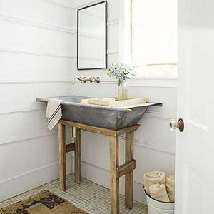 May include: A rustic bathroom sink made from a galvanized metal tub, set on a wooden stand. The sink has a towel draped over the edge and a wooden tray with items on it. The room has white walls, a mirror, and a small rug.