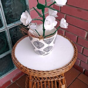 May include: White paper flowers in a white ceramic pot with a blue and white leaf pattern, sitting on a brown wicker table with a white top.