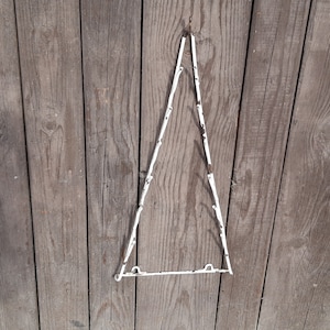 Può includere: Un appendino triangolare da parete in metallo bianco su uno sfondo di legno stagionato. Il triangolo è costruito con sottili aste collegate, con un gancio in alto per appendere e una barra orizzontale in basso.