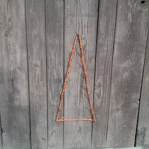 May include: An orange, triangular-shaped coat rack with multiple small pegs for hanging items. The rack is suspended against a weathered, gray wooden door. The design is simple and functional, suitable for a rustic or minimalist aesthetic.