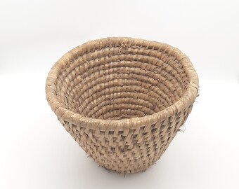 Puede incluir: Una cesta tejida de materiales naturales, con forma redonda y de cuenco. La cesta es de color marrón claro y tiene una textura ligeramente rugosa.
