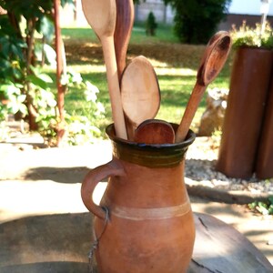 May include: A brown clay pitcher with a green rim holds a collection of wooden spoons. The spoons are various sizes and shapes, with some having a rounded end and others having a more pointed end.