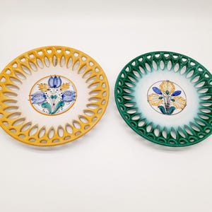 Op de afbeelding: Twee decoratieve keramische borden met een witte achtergrond en ingewikkelde uitgesneden ontwerpen. Een bord heeft een gele rand en de andere een groene rand. Beide borden hebben handgeschilderde bloemmotieven in blauw, geel en groen.