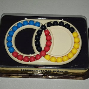 Peut inclure: Un jeu de puzzle hongrois "Hungarian Rings" dans une boîte noire et dorée. Le puzzle se compose de deux anneaux imbriqués avec des perles colorées.
