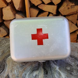 May include: A silver first aid kit with a red cross on the lid. The rectangular metal box has rounded corners and shows signs of wear. The background features a stack of logs.