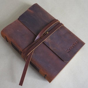 May include: A dark brown leather journal with a wrap-around strap closure and a small brass stud. The name Kristen is embossed on the front cover. The journal features visible stitched binding along the spine and a distressed, matte texture.