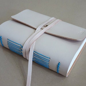 May include: A light beige leather journal with a flap closure and a wrap-around strap. The journal's spine is hand-stitched with bright blue thread. The journal is closed and has a small embossed logo on the bottom right corner.