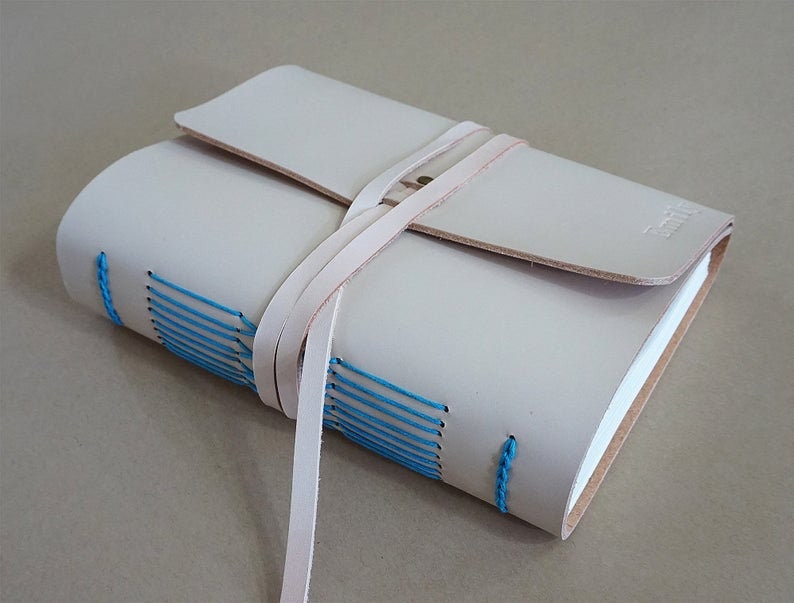 May include: A light beige leather journal with a flap closure and a wrap-around strap. The journal's spine is hand-stitched with bright blue thread. The journal is closed and has a small embossed logo on the bottom right corner.